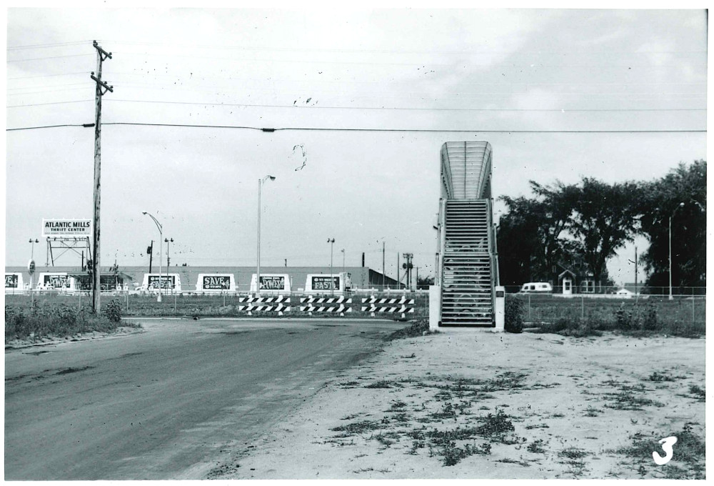 Black-and-white photo of the bridge taken from the ground straight on.  On the far side of the bridge we can see a building and a sign that says 'Atlantic Mills Thrift Center.'